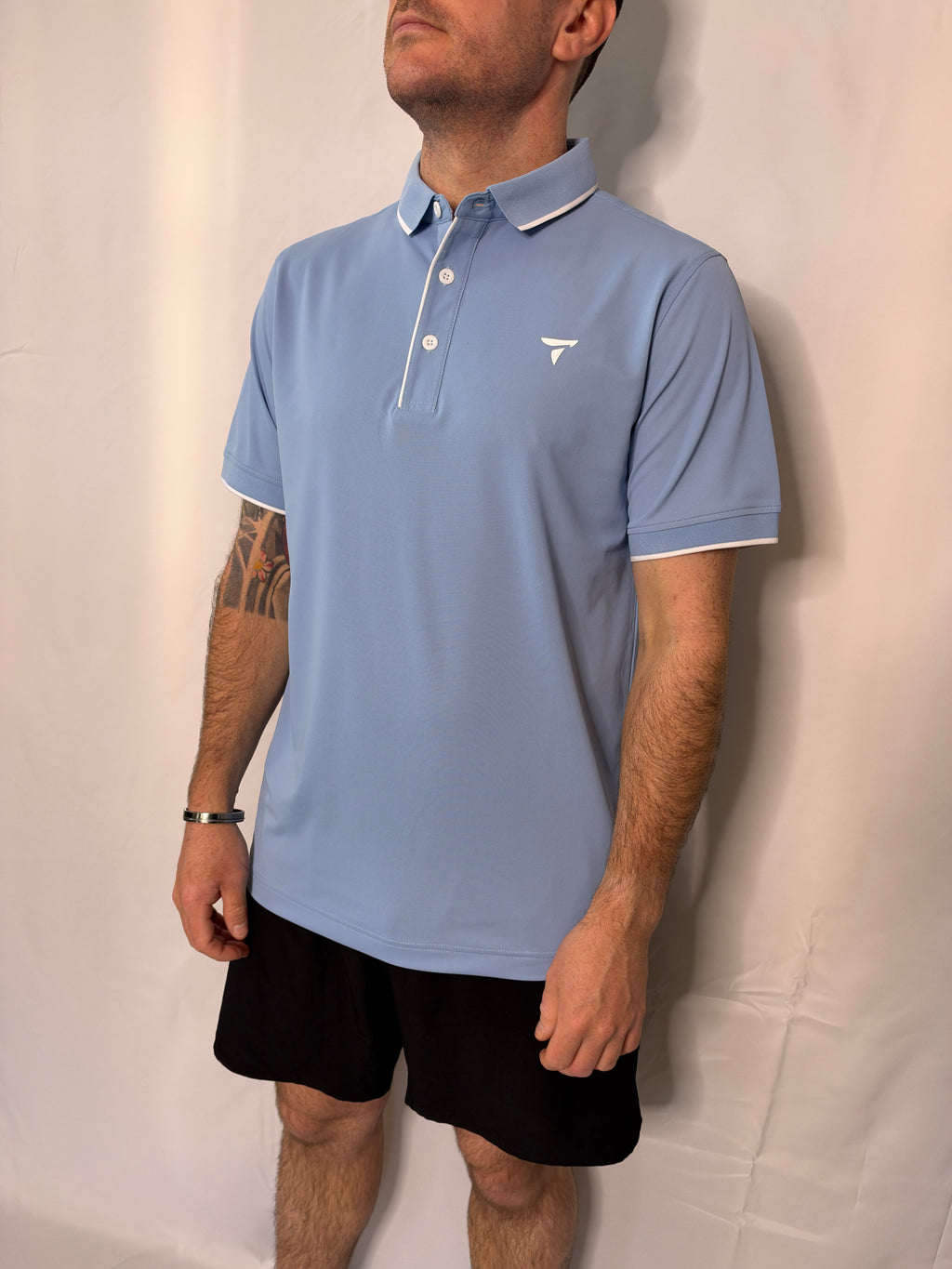 A man wearing a light blue short-sleeve polo shirt with white piping and a small white chest logo, paired with black shorts